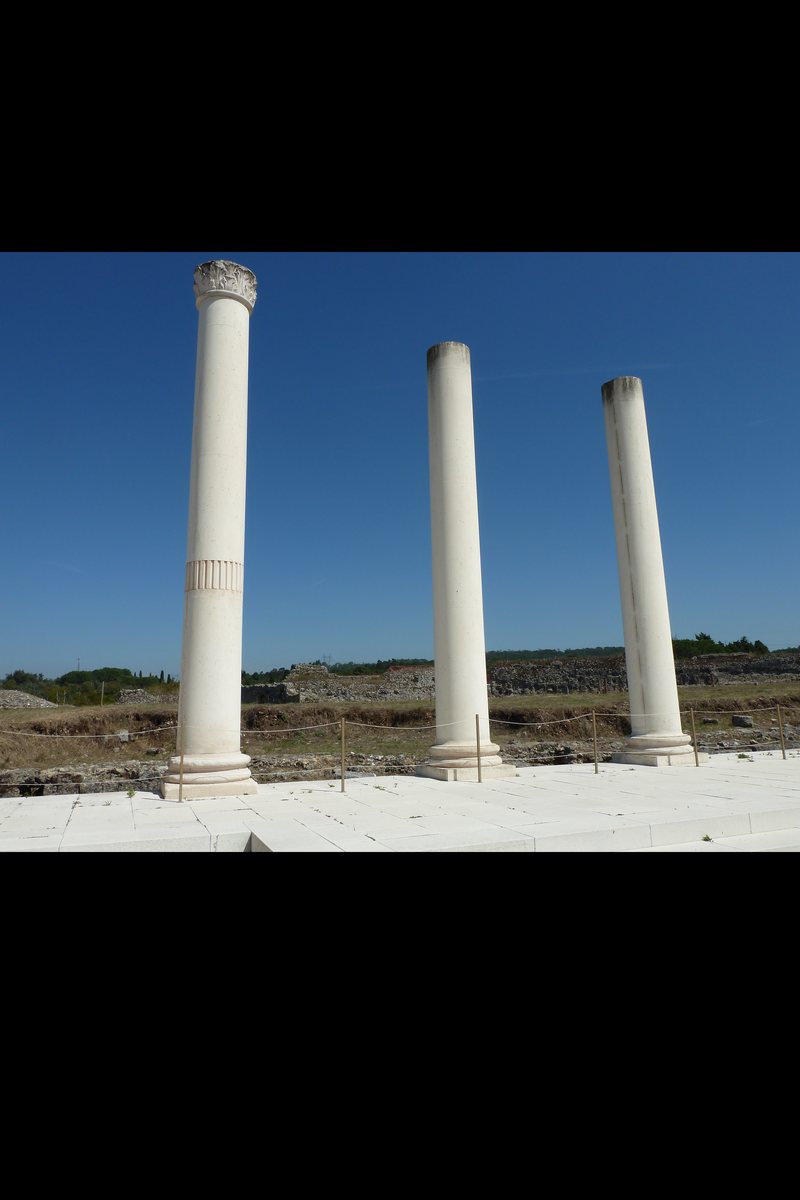 Reconstructed Corinthian columns standing over the excavated Roman town of Conímbriga, central Portugal, with the town's ruined stone walls stretching behind under a deep blue sky. One of Portugal's largest Roman archaeological sites, a National Monument since 1910.